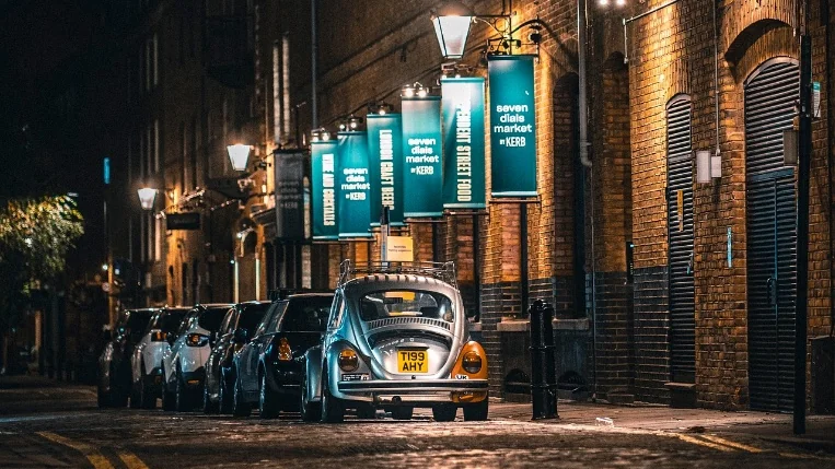 Central London side street at night with glowing venue banners, lamp light and a classic car parked by the kerb