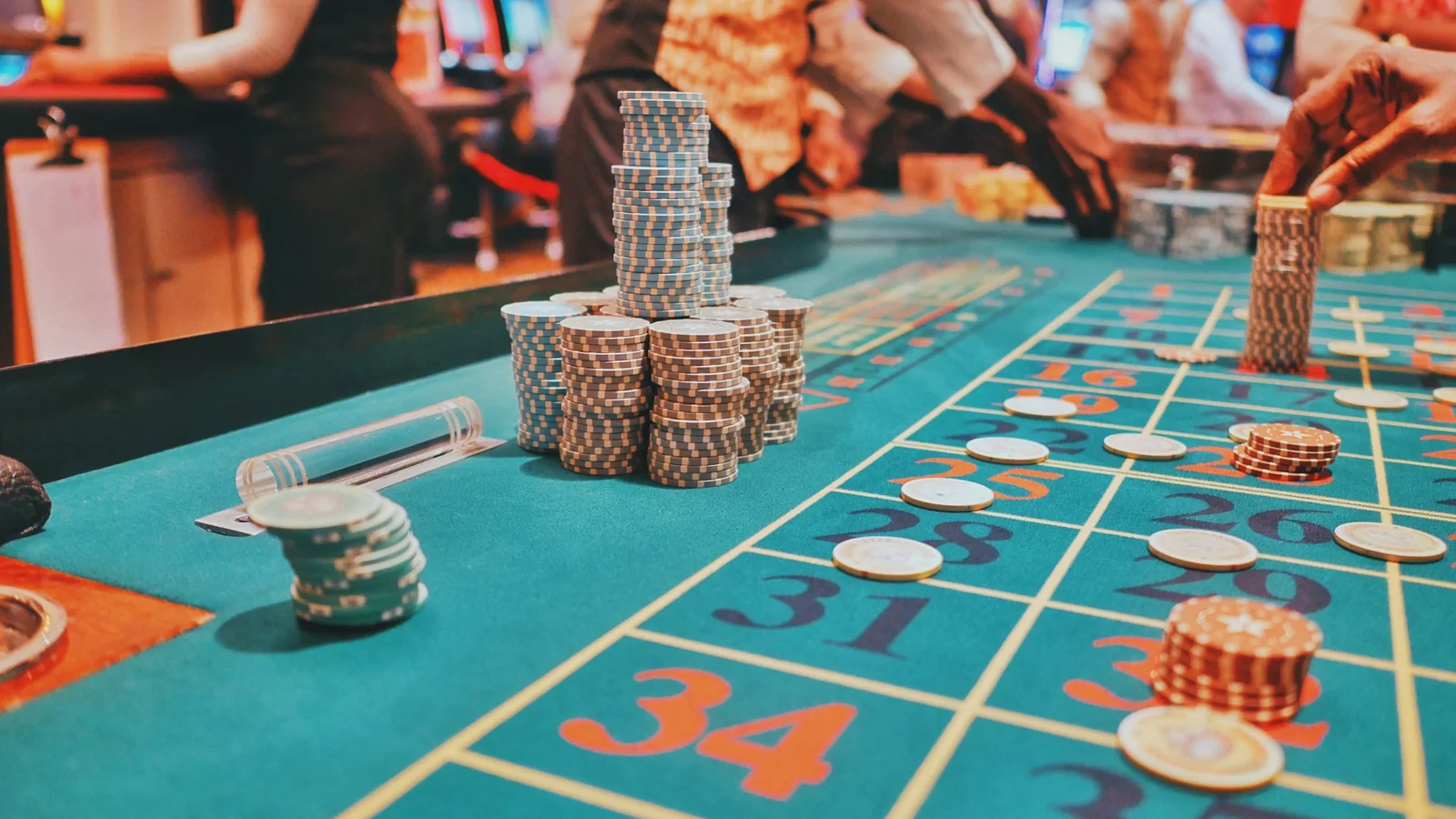 Stacked chips and placed bets on the numbered layout of a roulette table, with the dealer and players behind
