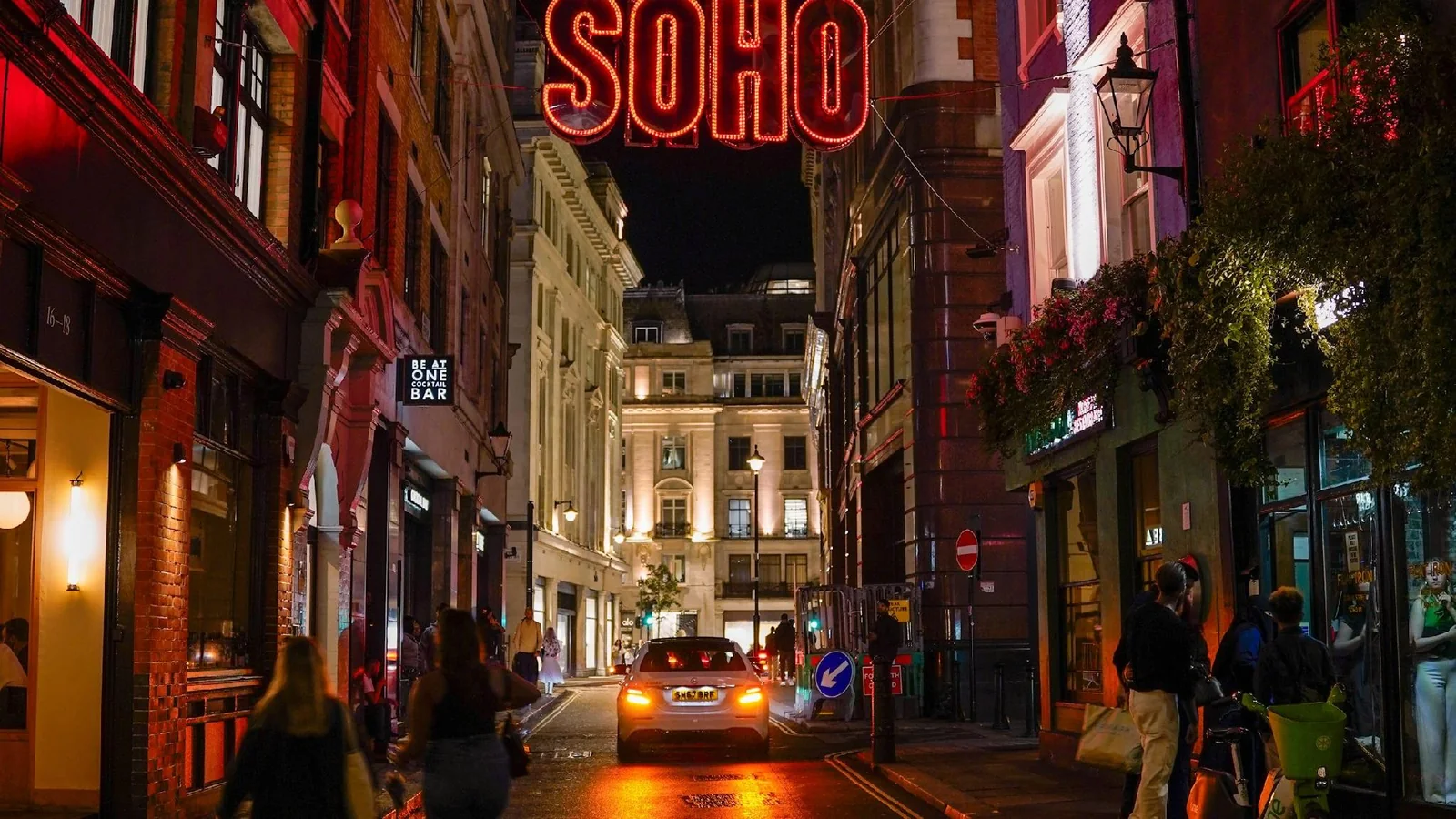 A Soho street at night beneath the red SOHO light sign, lined with bars and restaurants as people and a taxi pass through