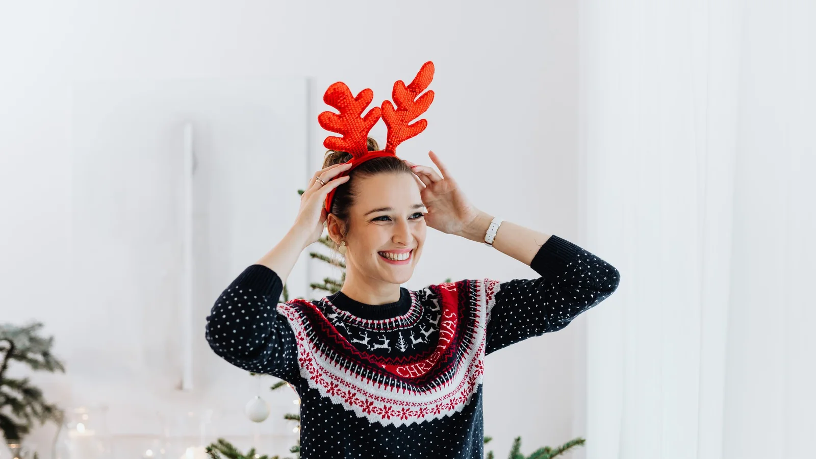 Woman in a navy patterned Christmas jumper adjusting a reindeer antler headband beside a decorated tree