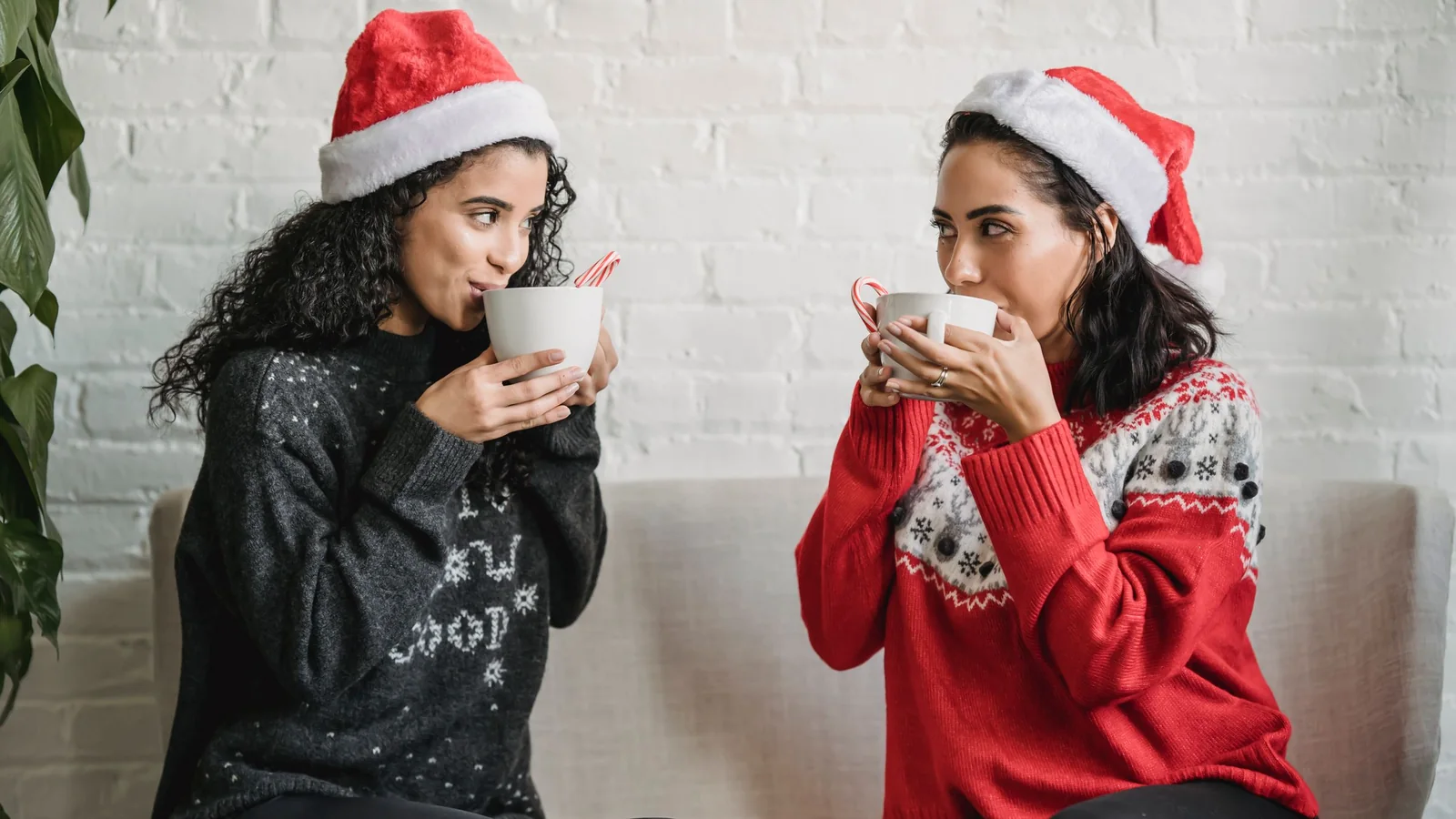 Two friends in Santa hats and Christmas jumpers sitting on a sofa drinking hot chocolate with candy canes
