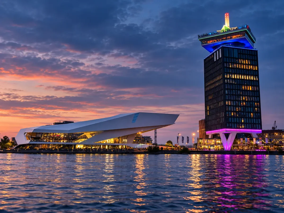 The EYE Filmmuseum and the A'DAM Tower lit against an orange and blue dusk sky on the north bank of the IJ in Amsterdam, their lights reflected in the water