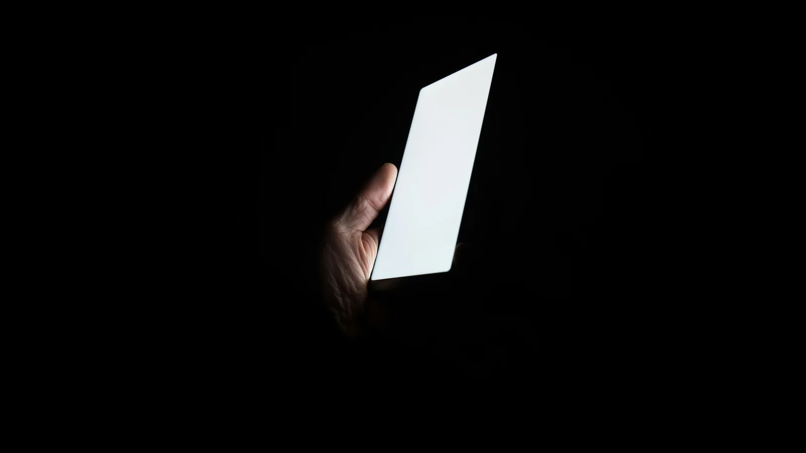 A hand holding a phone with a brightly lit blank screen, the only light source in an otherwise dark frame