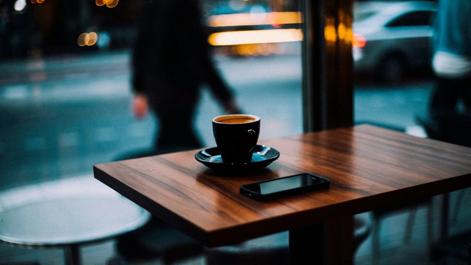 An espresso cup and a smartphone resting on a wooden cafe table beside a window onto a lit street