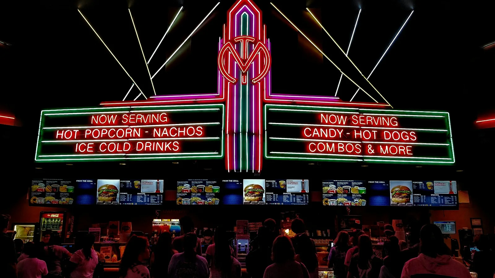 Cinemagoers queuing at a concession stand under a neon sign advertising hot popcorn, nachos, candy and cold drinks