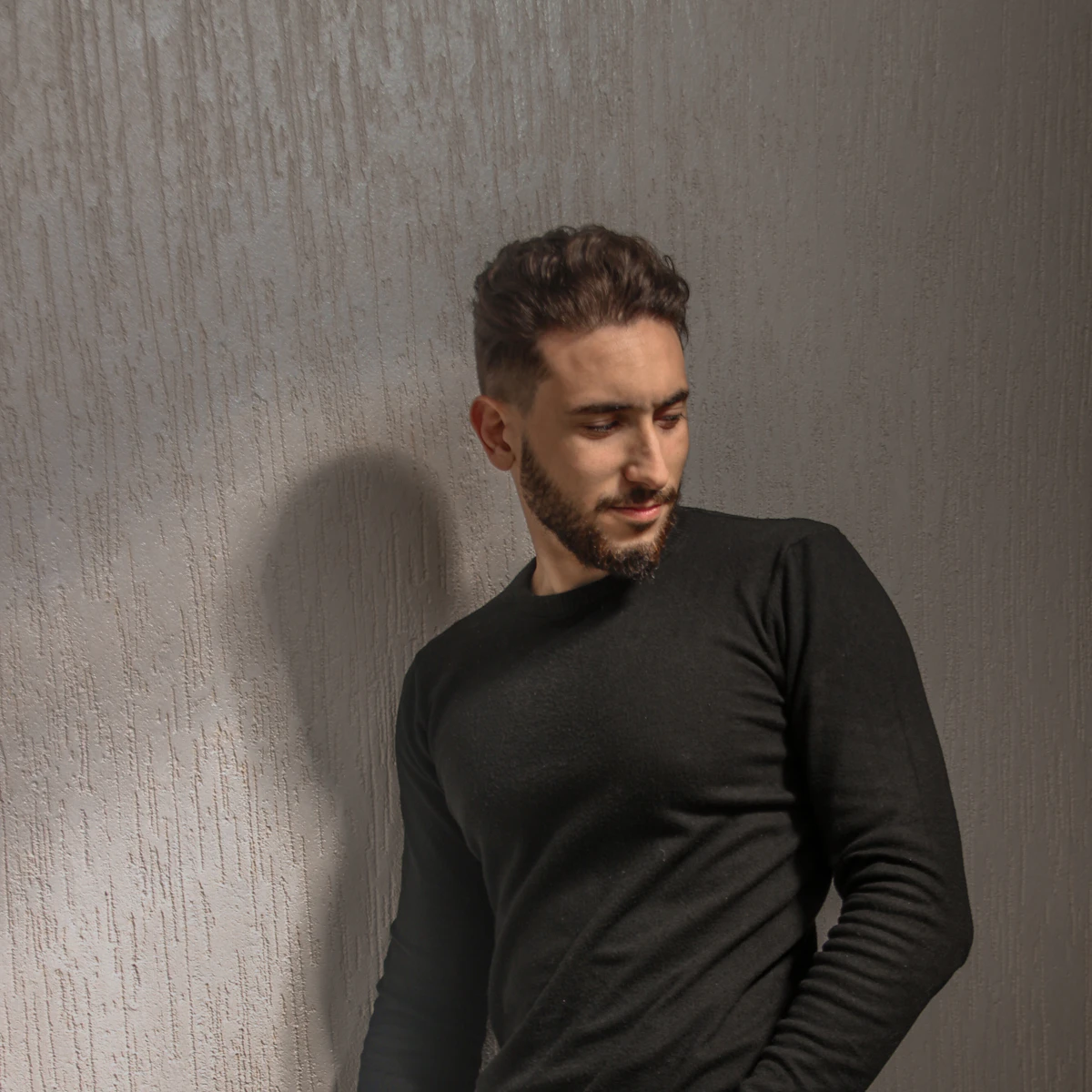 A man in a fitted black crewneck sweater leaning against a textured wall