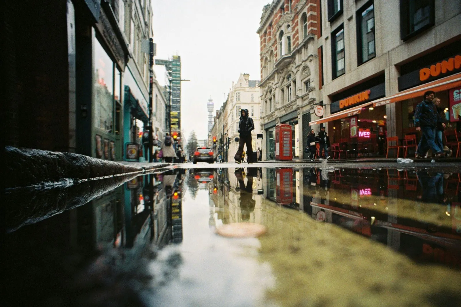 A London street after rain with buildings and a red telephone box reflected in a puddle and the BT Tower in the distance