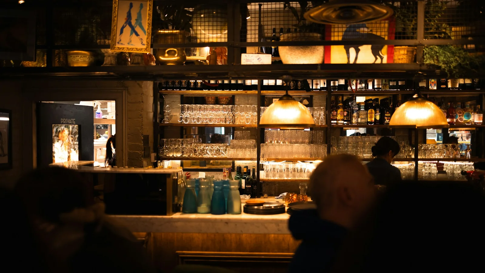 A dimly lit bar interior with bottles and glassware on shelves behind the counter and drinkers seated in the foreground