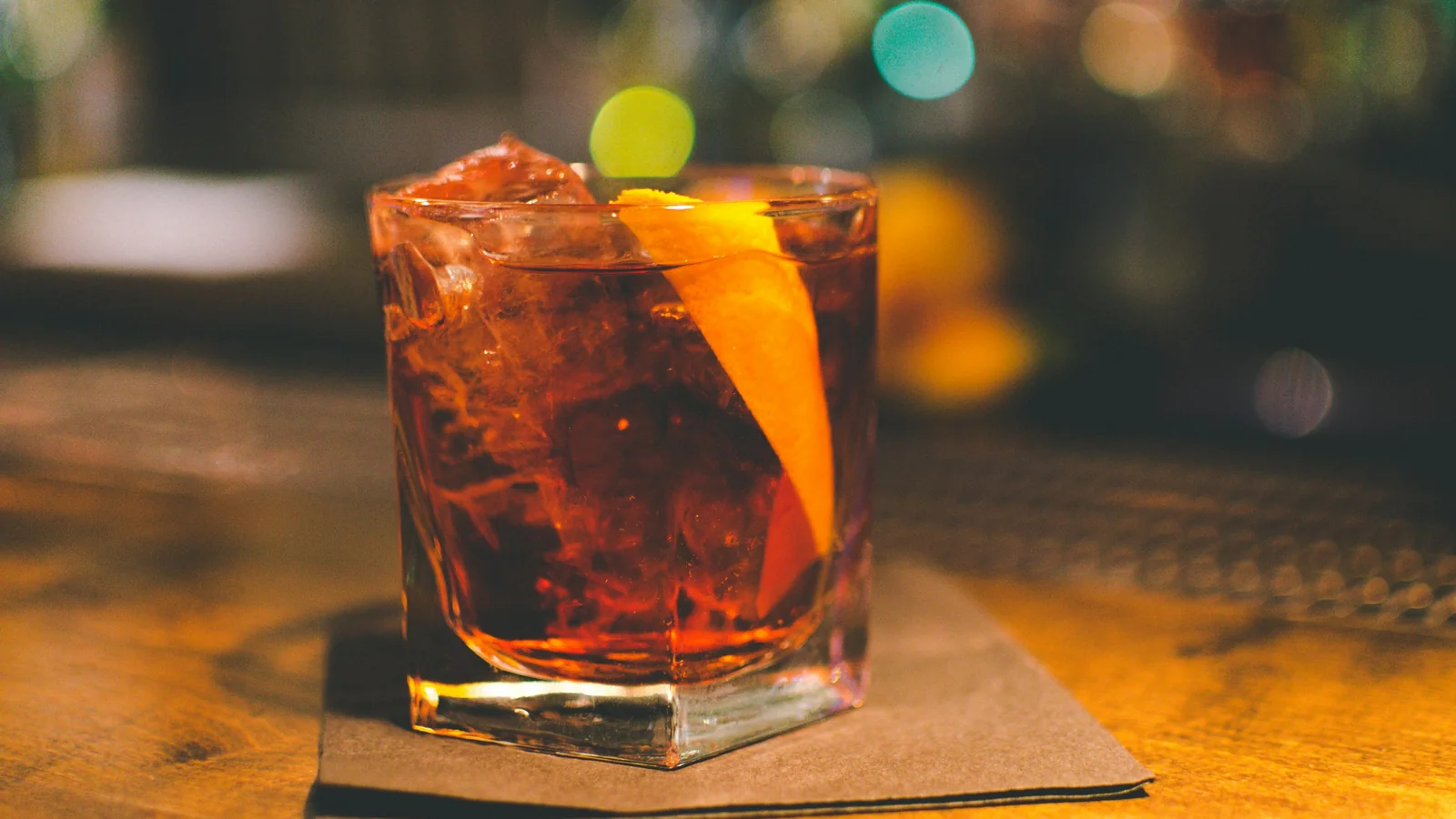 A Negroni served over ice with an orange peel garnish on a wooden bar top, with warm bar lights blurred behind