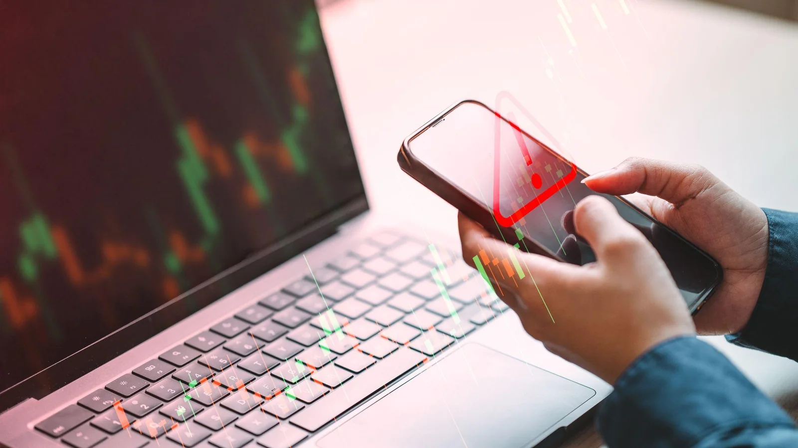 A laptop showing a candlestick chart beside a hand holding a phone displaying a falling price alert