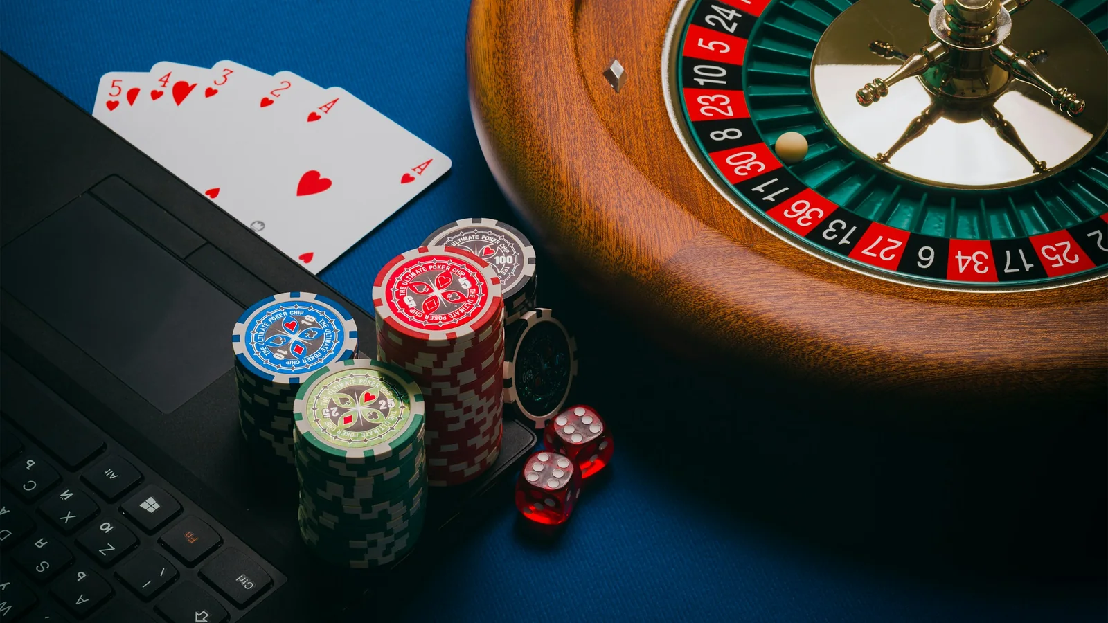 A roulette wheel, stacked poker chips, playing cards and dice arranged beside an open laptop keyboard